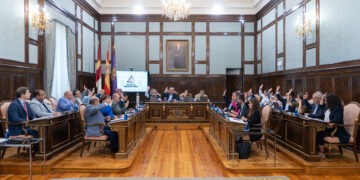 Los tramos de las carreteras provinciales GU-193 y GU-103 que discurren por los cascos urbanos de El Casar y de Azuqueca de Henares, respectivamente, pasarán a ser de titularidad municipal. El pleno de la Diputación de Guadalajara, actualmente titular de la totalidad de los trazados de ambas carreteras, ha aprobado en pleno las mutaciones demaniales subjetivas de ambas travesías a favor de los ayuntamientos de El Casar y de Azuqueca de Henares. El acuerdo del pleno de la Diputación se ha adoptado por unanimidad de todos los grupos políticos y responde a la voluntad expresada por ambos ayuntamientos de hacerse cargo de la titularidad de los tramos viales o travesías que pasan por sus respectivos cascos urbanos. Tanto el Ayuntamiento de El Casar como el de Azuqueca de Henares deberán ahora ratificar por acuerdo de sus respectivos plenos municipales la aceptación del cambio de titularidad del vial que afecta a cada uno de ellos para posteriormente firmar sendos convenios con la Diputación de Guadalajara. Cada uno de estos convenios establece que la Diputación ejecutará las actuaciones de adecuación necesarias para la transformación de cada tramo en vial urbano y, a partir de entonces, el Ayuntamiento asume todas las competencias inherentes a la conservación, mantenimiento, explotación, disciplina y gestión del tramo cedido. Para la transformación de los tramos en viales urbanos, los ayuntamientos deberán facilitar la disponibilidad de los espacios necesarios para la ejecución de las actuaciones y ejecutar previamente las canalizaciones o infraestructuras que resulten necesarias antes de las obras de acondicionamiento. 1,3 km y 9.114 metros cuadrados afectados en El Casar. La carretera GU-193 es la vía provincial que va desde la carretera N-320 hasta el límite provincial por El Casar. El tramo cuya titularidad va a pasar de provincial a municipal es el que comprende el recorrido existente desde la intersección con la carretera N-320 hasta el inicio de la rotonda de acceso al centro de salud de la localidad. Este tramo tiene una longitud total de 1.323,70 metros y una superficie de 9.114 metros cuadrados. El servicio de Infraestructuras de la Diputación ha redactado el proyecto de actuación para acondicionar este tramo como vial urbano, en el que se definen técnica y económicamente las obras de adecuación que la propia Institución Provincial llevará a cabo. Dichas actuaciones para adaptar el tramo a su nueva funcionalidad urbana consistirán en: fresado y extendido de nueva capa de rodadura, repintado de marcas y símbolos viales y cerramiento de la rotonda del centro de salud mediante bordillo, capa de adoquín perimetral, acabado interior de rocalla, colocación de dos puntos de luz, plantación de dos árboles, instalación de captafaros LED solares en bordillos interiores y mejora de señalización de seis pasos de peatones. 1,15 km y 20.072,74 metros cuadrados en Azuqueca. En el caso de Azuqueca de Henares, la carretera provincial GU-103 es la que sale desde la GU-102 y lleva al límite de la provincia de Guadalajara y a Meco. El tramo cuya titularidad cede la Diputación al Ayuntamiento azudense es el comprendido entre la Avenida de Alcalá y el cruce con la calle Rafael Guijosa de la localidad, mientras que el tramo situado desde ese punto hasta el límite de la provincia seguirá siendo de titularidad provincial. Al igual que en El Casar, la Diputación se hará cargo de la adaptación del tramo afectado como vial urbano ejecutando el proyecto que ha elaborado el servicio provincial de Infraestructuras. De esta forma, en los 1.156,12 metros de longitud y 20.072 metros cuadrados de superficie que pasan a ser de titularidad municipal, la Diputación llevará a cabo las siguientes actuaciones: fresado y extendido de nueva capa de rodadura, repintado de marcas y símbolos viales, adecuación del interior de la rotonda de enlace con la calle Rafael Guijosa mediante cerco de adoquín, relleno del interior con sistema drenante y marmolina blanca, plantación de magnolio y señalización de todas las rotondas cedidas con catadióptricos leds solares.